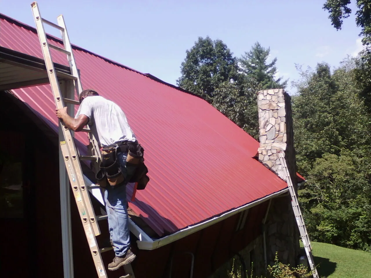 Roofer on a ladder beside a red metal roof and stone chimney — Soffit & Fascia Repair by Roofing Brothers in Greenbackville